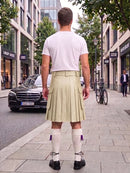 A man viewed from behind wearing a white t-shirt and a light beige pleated kilt with white knee-high socks, standing on a city sidewalk.