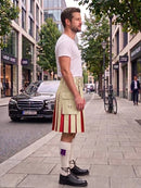 A man standing on a city sidewalk wearing a white kilt with red and white pleated detailing, a white shirt, white socks, and black shoes, with buildings, trees, and parked cars in the background.