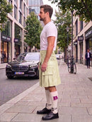 A man standing on a city sidewalk wearing a pale yellow-green pleated kilt with embroidered details, a white t-shirt, white socks with a purple bow, and black shoes. A black Mercedes-Benz car and urban buildings are visible in the background.