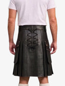A man wearing a black faux-leather kilt with a lace-up back, side pockets, and pleated front, paired with a white t-shirt and white socks, shown from the back against a white background.