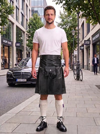 A man stands on a city sidewalk wearing a black leather kilt with a front pouch, a white crew-neck t-shirt, white knee-high socks, and black leather shoes. Buildings, trees, and parked cars are visible in the background.