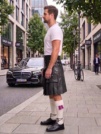 A man standing on a city sidewalk wearing a black leather kilt, white t-shirt, white socks with a purple accent, and black leather shoes. A black Mercedes-Benz car is parked on the street behind him.
