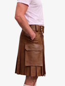 A man wearing a brown leather kilt with pleats, a belt, and a large front flap pocket, paired with a light pink t-shirt, shown from the waist down against a plain white background.