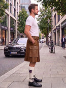 A man standing on a city sidewalk wearing a brown leather kilt, white t-shirt, white socks, and black leather shoes, with a street scene in the background.