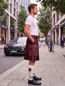 A man standing on a city sidewalk wearing a dark green pleated kilt, a white t-shirt, white socks, and black leather shoes. A black Mercedes-Benz car is parked on the street behind him, with trees and storefronts in the background.