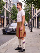 A man stands on a city sidewalk wearing a tan pleated kilt with red and gold accents, a white t-shirt, white knee-high socks, and black leather shoes. A black Mercedes-Benz car and storefronts are visible in the background.