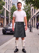 A man stands on a city sidewalk wearing a white short-sleeve t-shirt and a dark gray faux-leather kilt with a matching belt and silver buckle. He is also wearing white knee-high socks and black leather shoes.
