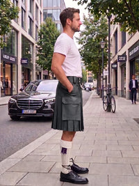 A man standing on a city sidewalk wearing a dark green pleated kilt, a white t-shirt, white socks, and black leather shoes. A black Mercedes-Benz car is parked on the street behind him, with trees and storefronts in the background.