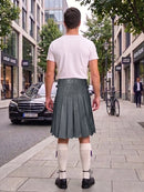 A man standing on a city sidewalk, viewed from behind, wearing a white t-shirt, a dark gray pleated leather kilt, white knee-high socks, and black shoes. Buildings, trees, a black Mercedes-Benz, and a bicycle are visible in the background.