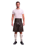 A man modeling a black faux-leather kilt with a matching black waist belt and pouch, paired with a white t-shirt, cream knee-high socks, and black lace-up shoes, against a plain white background.