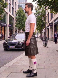 A man standing on a city sidewalk wearing a dark brown leather kilt, white t-shirt, white socks, and black shoes, with a black Mercedes-Benz car and urban buildings in the background.