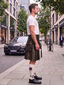 A man standing on a city sidewalk wearing a black leather kilt, white t-shirt, white knee-high socks, and black leather shoes. A black Mercedes-Benz car is parked in the background on a city street.