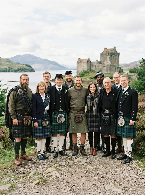 A group of ten people posing outdoors in traditional Scottish attire, including kilts and sporranes, with a scenic view of a castle and lake in the background.