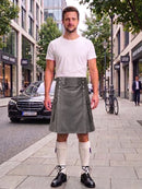 A man stands on a city sidewalk wearing a white crew-neck t-shirt and a gray pleated kilt with silver metal accents, paired with white knee-high socks and black leather shoes. A black Mercedes-Benz car and a bicycle are visible in the background of the street scene.