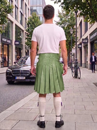 A man standing on a city sidewalk, photographed from behind, wearing a white t-shirt, a green pleated leather kilt, white knee-high socks, and black shoes. A black Mercedes-Benz car is parked on the street to his left, with storefronts and trees in the background.