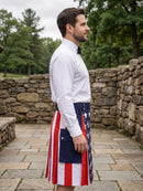 A man in a white dress shirt and black bow tie stands in profile outdoors wearing a kilt with an American flag pattern featuring red and white stripes and a blue panel with white stars.