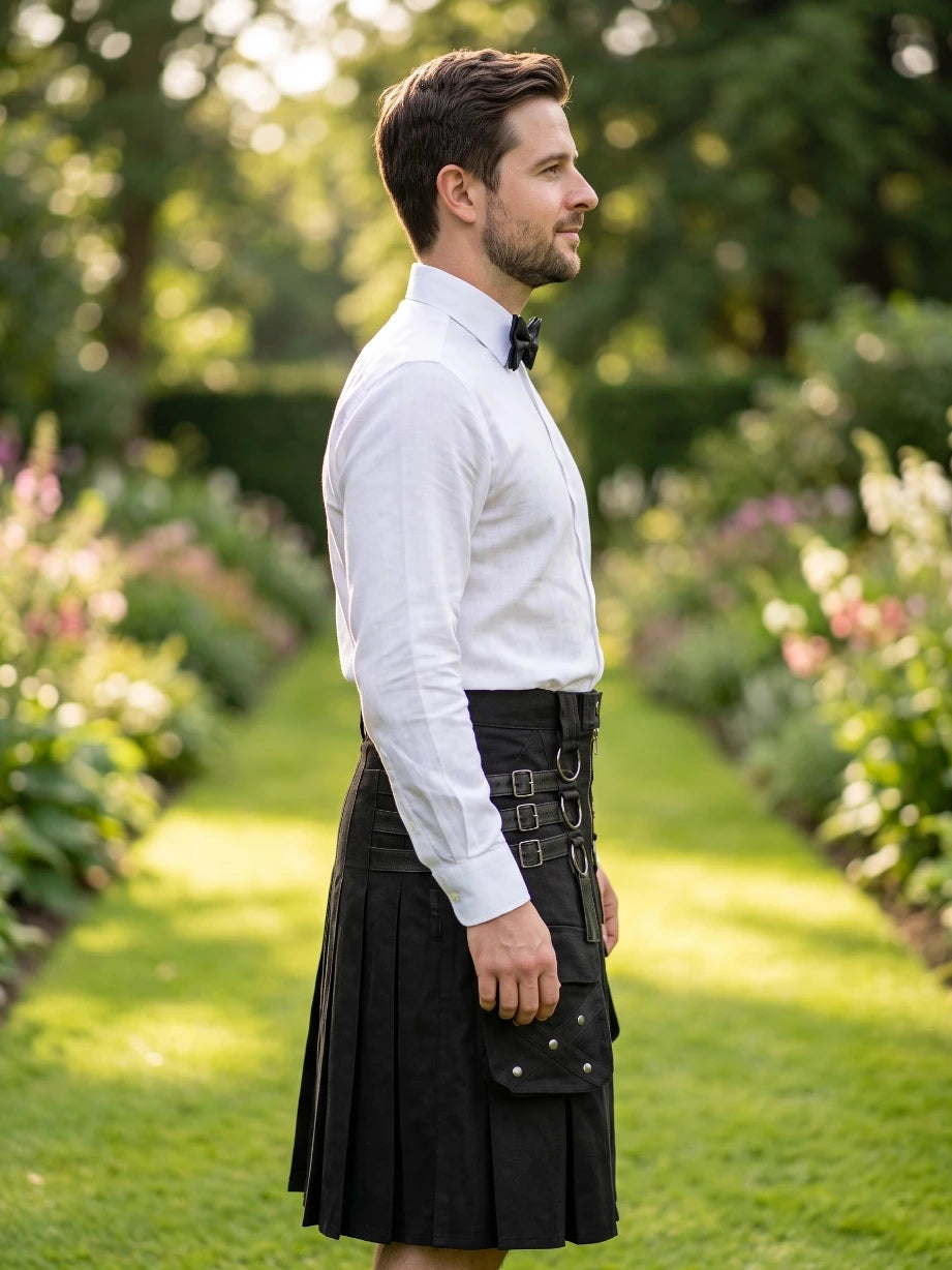 A man standing in profile in a sunlit garden path, wearing a white long-sleeve dress shirt, black bow tie, and a black kilt with a front flap pocket.