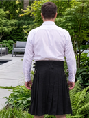 A man viewed from behind wearing a white dress shirt and a black pleated kilt in an outdoor garden setting.