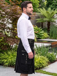A man wearing a black kilt with silver hardware, a white dress shirt, and a black bow tie, standing in a landscaped garden with plants and a water feature in the background.