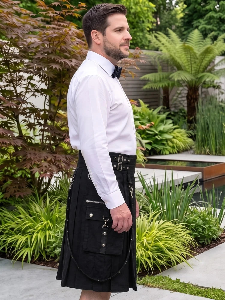 A man wearing a black kilt with silver hardware, a white dress shirt, and a black bow tie, standing in a landscaped garden with plants and a water feature in the background.