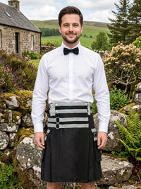 A man wearing a white dress shirt, black bow tie, and a black kilt with a wide gray and black striped waist belt, standing outdoors in front of a stone wall and green hills.