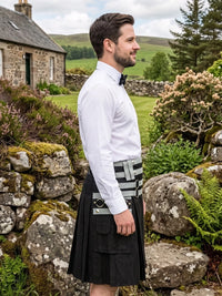 A man in profile wearing a white long-sleeve dress shirt, black bow tie, and a black kilt with a gray and black striped waist belt, standing outdoors in a rural Scottish landscape with stone walls, green hills, and a cottage in the background.