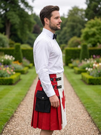 A man standing in a formal garden wearing a white long-sleeve dress shirt, black bow tie, and a red and white kilt with black belt and pouch details.