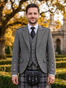 A man wearing a formal Scottish outfit with a gray herringbone blazer, matching vest and tie, a plaid kilt, and a black sporran bag, standing outdoors in front of a blurred garden and stone building.