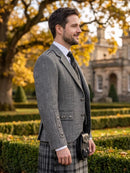 A man in profile wearing a gray herringbone blazer, white dress shirt, dark tie, and a gray plaid kilt with a sporran, standing outdoors in front of a stone building and autumn trees.