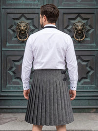 A man viewed from behind wearing a white long-sleeve dress shirt and a gray pleated herringbone kilt, standing in front of a large dark green ornate door with lion head knobs.