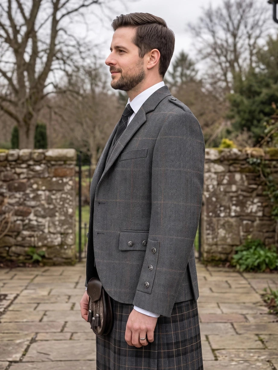 A man in profile wearing a gray windowpane plaid blazer, a dark plaid kilt, a white dress shirt, and a black tie, holding a black sporran outdoors in front of a stone wall.