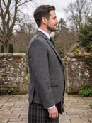 A man in profile wearing a gray windowpane plaid blazer over a white shirt and tie, paired with a dark plaid kilt, standing outdoors in front of a stone wall.