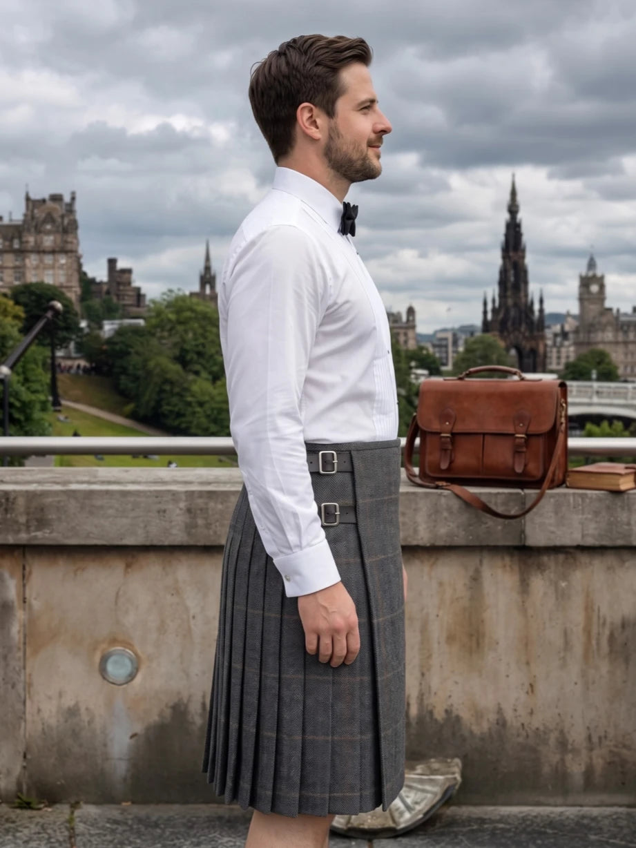 A man in profile wearing a white dress shirt, black bow tie, and gray plaid kilt, standing outdoors on a stone bridge with Edinburgh castle and spires in the background. A brown leather satchel rests on the wall beside him.