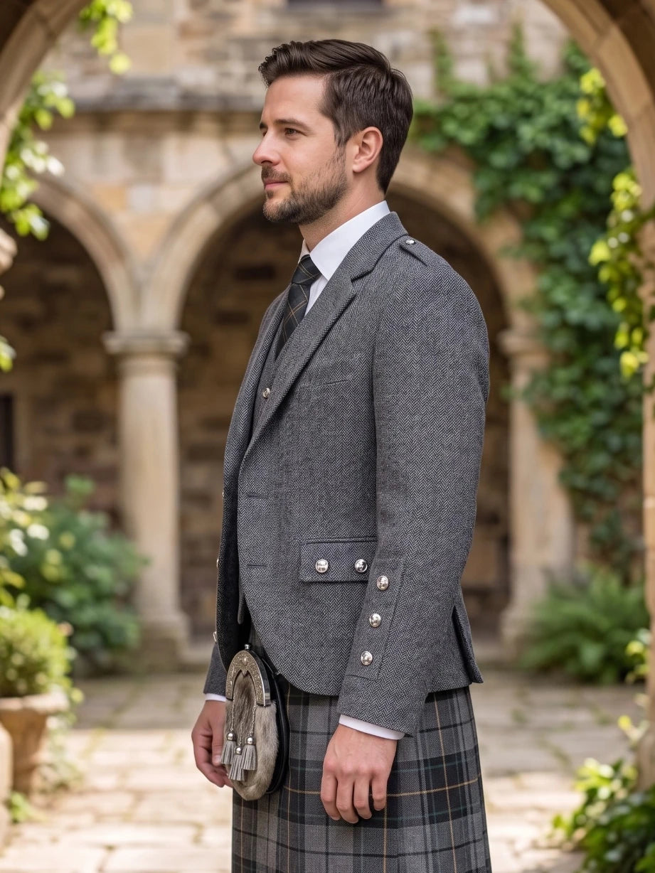 A man in profile wearing a gray tweed blazer, white dress shirt, dark tie, and a gray plaid kilt, standing outdoors in front of a stone courtyard with arches and greenery.