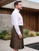 A man wearing a brown pleated kilt with front flap pockets, styled with a white dress shirt and black bow tie, standing outdoors in front of a modern wooden building.