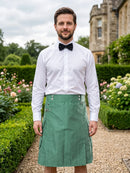 A man wearing a white dress shirt, black bow tie, and a green kilt stands in a garden setting with hedges and a stone building in the background.