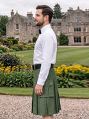 A man wearing a white dress shirt, black bow tie, and an olive green kilt with a black belt, standing in a gravel path with a large stone mansion and green lawn in the background.