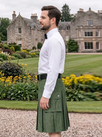 A man wearing a white dress shirt, black bow tie, and an olive green kilt with a black belt, standing in a gravel path with a large stone mansion and green lawn in the background.