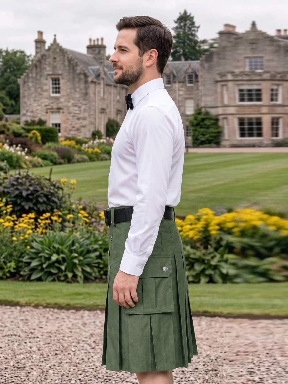A man wearing a white dress shirt, black bow tie, and an olive green kilt with a black belt, standing in a gravel path with a large stone mansion and green lawn in the background.