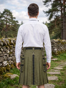 A man viewed from behind wearing a white long-sleeve dress shirt and an olive green pleated kilt with black trim and side cargo pockets, standing outdoors on a stone path with trees and a stone wall in the background.