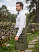 A man wearing a white dress shirt, black bow tie, and an olive green kilt with black straps and brass hardware, standing outdoors in a grassy path with a stone wall and pine trees in the background.