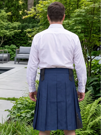 A man viewed from behind wearing a white dress shirt and a navy blue pleated kilt with black leather side tabs, standing in a lush green outdoor garden setting.