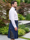 A man wearing a navy blue kilt with black leather trim and brass grommets, paired with a white dress shirt and black bow tie, standing in a landscaped garden.