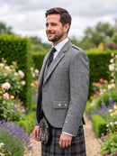 A man wearing a gray tweed blazer, white dress shirt, dark tie, and a black-and-gray plaid kilt with a sporran, standing in a lush garden pathway.