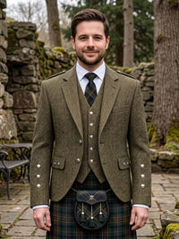 A man wearing a traditional Scottish outfit, featuring an olive green tweed blazer with silver buttons, a matching waistcoat, a white dress shirt, a plaid tie, and a tartan kilt with a sporran bag, standing outdoors in front of a stone wall and trees.