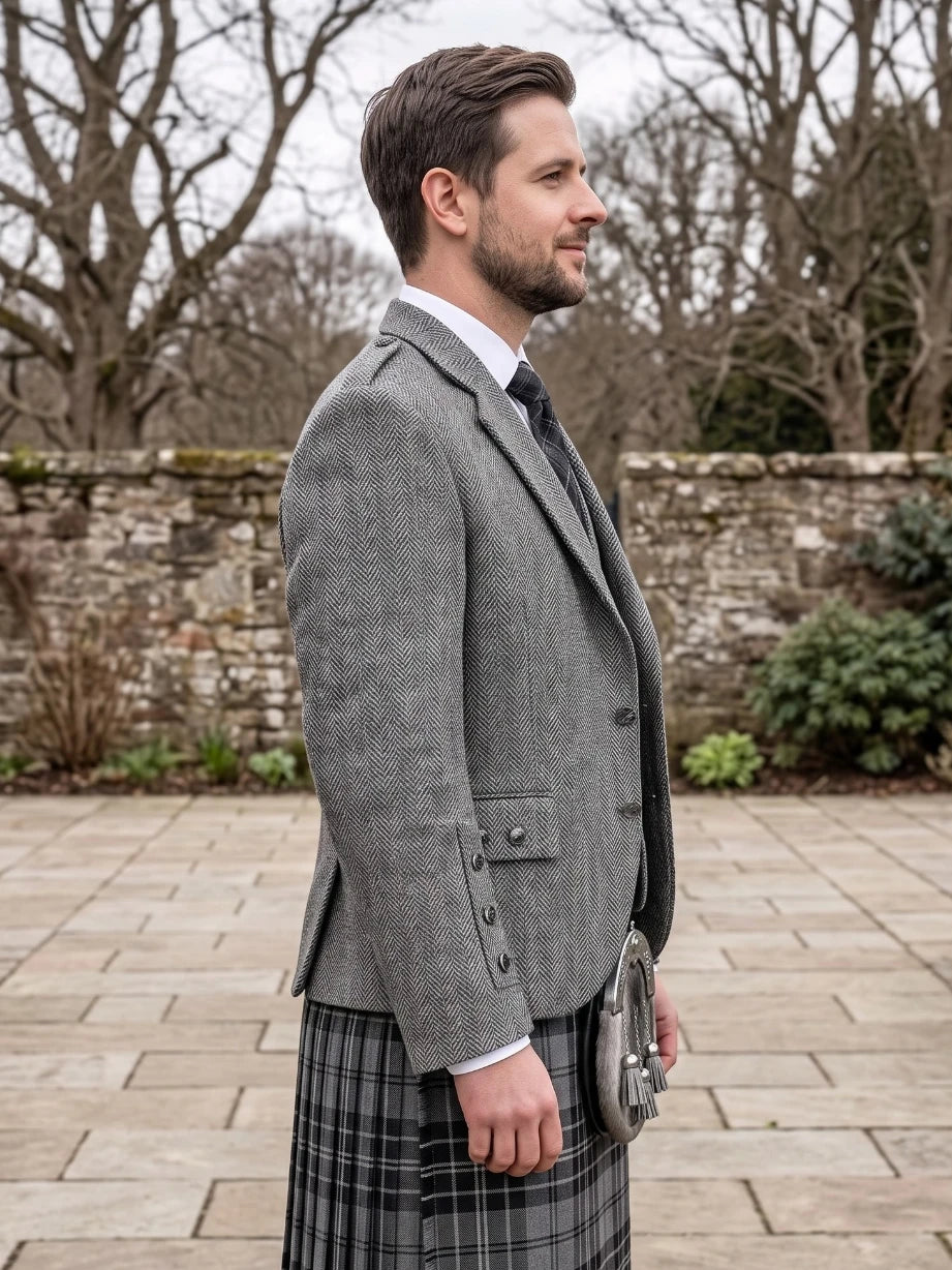 A man in profile wearing a gray herringbone tweed blazer, a black and gray plaid kilt, a white dress shirt, and a dark tie, standing outdoors on a stone paved area with a stone wall and bare trees in the background.