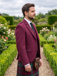 A man wearing a burgundy kilt jacket, white dress shirt, green patterned tie, and a tartan kilt stands in a garden setting.