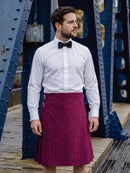 A man wearing a white dress shirt, black bow tie, and a magenta pleated kilt with silver buckle details, standing on a wooden walkway in front of a blue steel bridge.