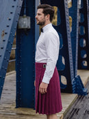 A man wearing a white long-sleeve dress shirt and a burgundy pleated kilt with a black bow tie, standing on a wooden walkway under a blue steel bridge.