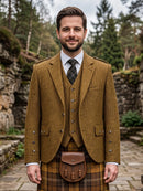 A man wearing a traditional Scottish outfit, featuring a brown corduroy blazer and waistcoat over a white shirt and dark plaid tie, paired with a brown leather sporran and a brown and yellow tartan kilt, set against a blurred outdoor background of stone walls and pine trees.